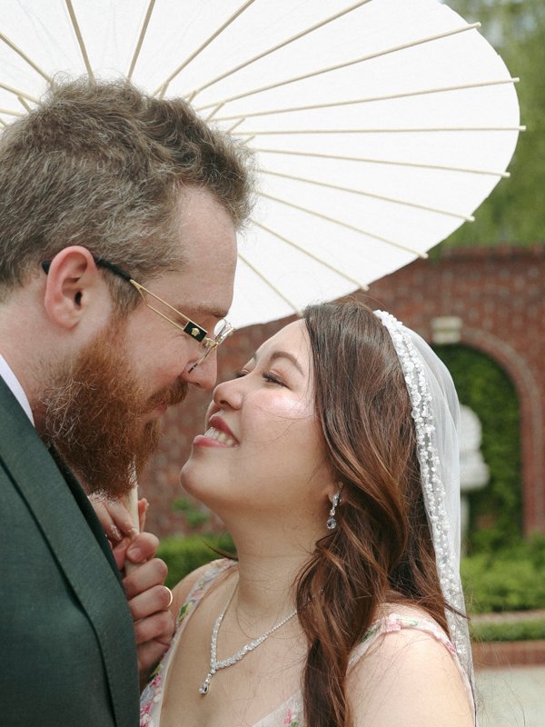 Bride and groom sharing a close, joyful moment under a white parasol during an outdoor wedding portrait session.